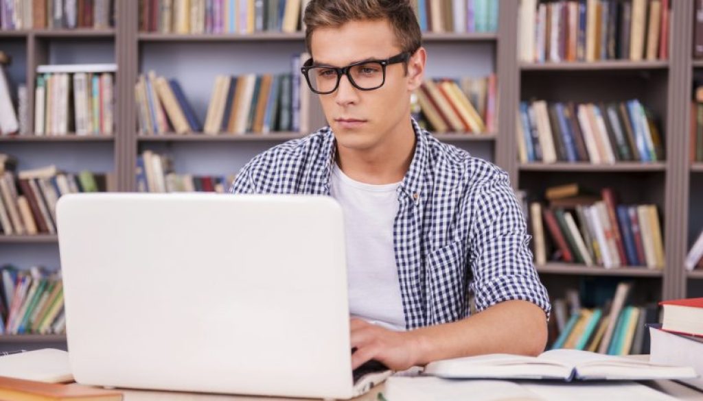 Preparing to exams in library. Confident young man working on laptop while sitting at the desk and in font of bookshelf